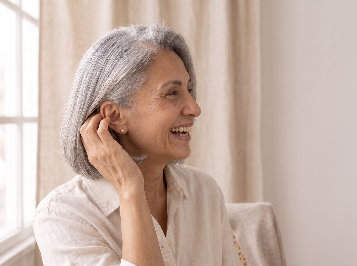 A person with a silver bob laughing and tucking a strand of hair behind one ear, by a window